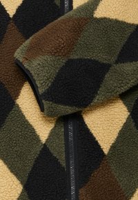 Καρό ύφασμα fleece σε πράσινο, καφέ, μαύρο και μπεζ με εμφανές μαύρο φερμουάρ και ζεστή, υφασμάτινη επιφάνεια.