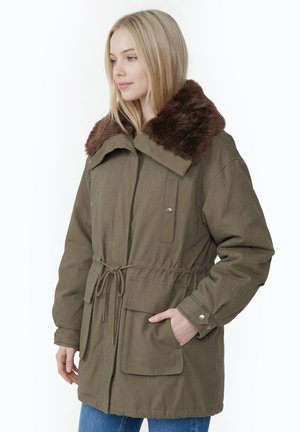 Junge Frau trägt eine olivgrüne Parka mit braunem Kunstpelzkragen und vorderen Taschen, steht mit einer Hand in der Tasche.