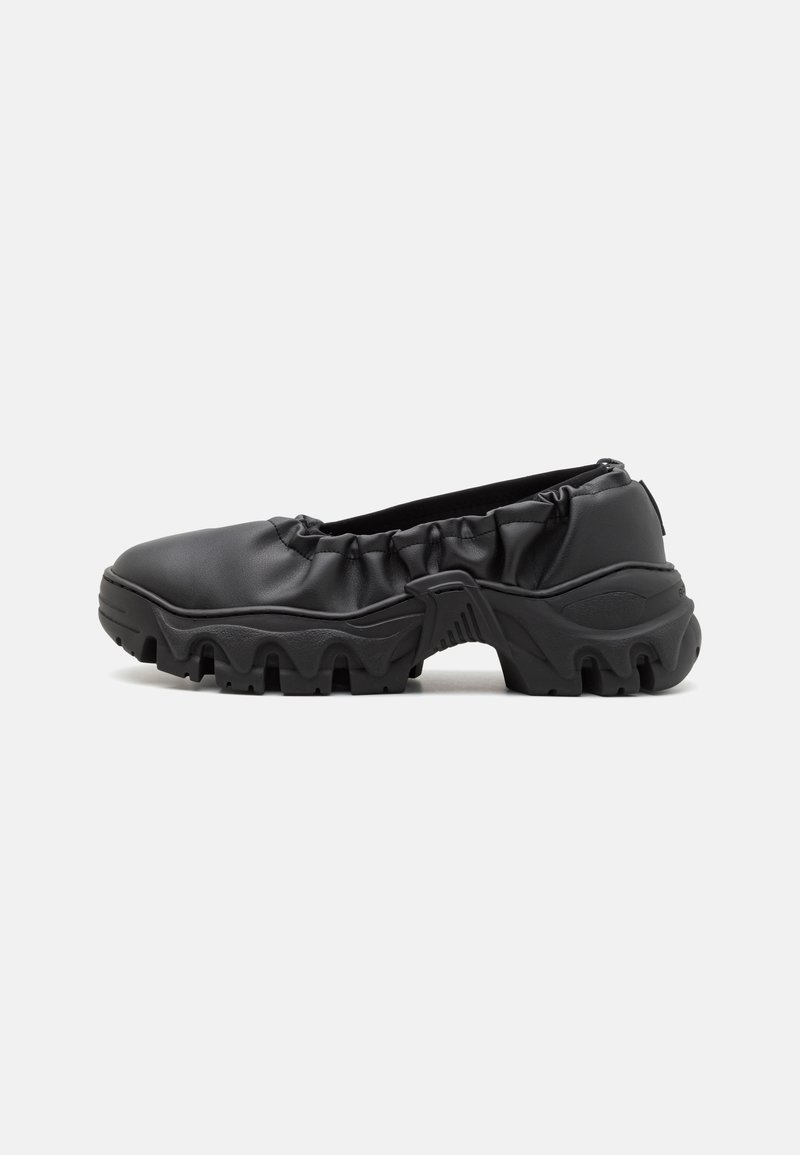 Black slip-on shoe made from smooth synthetic material. Features a gathered elasticized upper and a chunky, textured rubber sole.