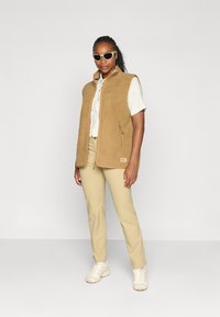 Brun fleece vest med en glidelåslomme, paret med en lys skjorte og beige bukser. Hvite klumpete joggesko og solbriller fullfører antrekket.