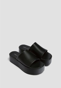 Des sandales à plateforme noires présentent un dessus en cuir lisse et brillant avec de larges brides, une semelle épaisse et un design minimaliste.