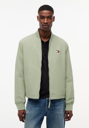 Fiatal férfi világoszöld cipzáras bomber dzsekiben, fekete galléros pólóval és kék farmerral, egyszerű fehér háttér előtt áll.