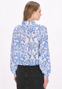 Bluse in hellblau mit floralem Muster, hohem Rüschen-Ausschnitt und weit schwingenden Ärmeln. Trägt sie über dunklen Jeans. Strukturierter Stoff.