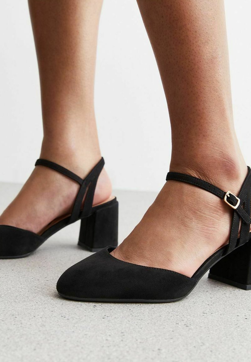 Scarpe a tacco blocco nere in suede con punta appuntita e cinturino alla caviglia con fibbia dorata, caratterizzate da una texture liscia e un design minimalista.