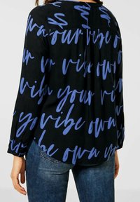 Svart blus med blå kalligrafisk text "your vibe own" övertygande på tyget. Långa ärmar och en avslappnad passform med en subtil veckning på baksidan.