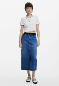 Denim maxi kjol i medelblå tvätt med rak silhuett, hög slits och svart elastisk midja. Kombineras med en figurnära vit topp.