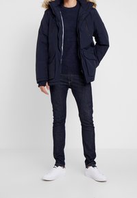 Giacca parca blu marina con cappuccio foderato in pelliccia rimovibile, chiusura zip, due tasche sul petto e due tasche laterali, indossata sopra un maglione blu marina e jeans scuri.