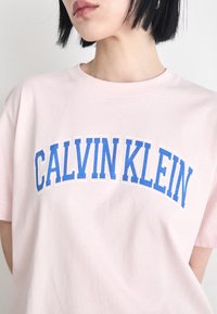 Rozā kokvilnas T-krekls ar uzrakstu "CALVIN KLEIN" zilās blokkavās uz krūšu daļas. Īsās piedurknes un apaļas kakla dizains.