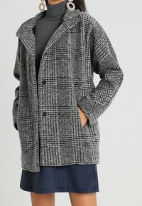 Grå och svart rutigt kappliknande plagg tillverkat av texturerad väv, med krage, två framfickor och knäppning.