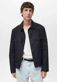 Zwarte, knopenjack met twee frontzakken, voorzien van een kraag, rechte snit en lichtgewicht, gestructureerde stof. Draagt over een wit shirt.