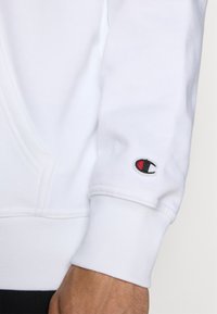 White sweatshirt with a soft texture, featuring a small embroidered logo in red and black on the cuff. Includes a kangaroo pocket.