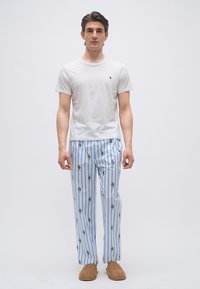 Άνδρας που φοράει λευκό μπλουζάκι, μπλε-λευκή ριγέ πιτζάμα με μικρά σχέδια πάντα και καφέ παντόφλες, στέκεται μπροστά σε απλό ανοιχτό φόντο.