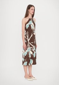 Robe halter-neck marron avec des motifs floraux bleus et blancs, longueur genou, en tissu léger, accompagnée de chaussures blanches à bout pointu.