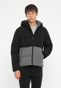 Sort og grå polstret jakke med hætte, med lynlås foran, to sidelommer og en struktureret finish. Bemærkelsesværdigt farveblokeringsdesign.