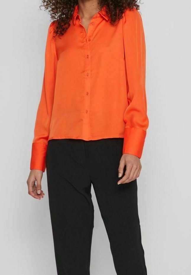 Chemise orange en satin avec boutons, manches longues, col et ourlet droit, associée à un pantalon noir.