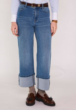 Persona che indossa jeans blu a gamba larga con risvolto, calzini grigi, mocassini marroni con nappine e una camicia a righe con braccialetti e anelli visibili.