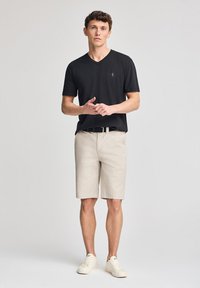 Camiseta negra de cuello en V, pantalones cortos beige y zapatillas blancas. El atuendo presenta un logo sutil en la camiseta y un cinturón negro con una hebilla plateada.