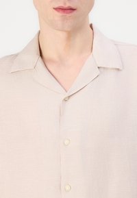 Camisa de linho beige clara com uma textura suave, colarinho curto e três botões na frente. Design simples e minimalista.
