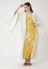 White textured kimono with yellow trim, layered over a yellow dress featuring white leaf patterns. Brown slip-on sandals complete the look.
