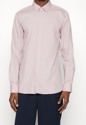 Mężczyzna ubrany w jasnoróżową, długorękawową koszulę zapinaną na guziki z kołnierzykiem w szpic oraz ciemnogranatowe spodnie, stojący na jednolitym tle.