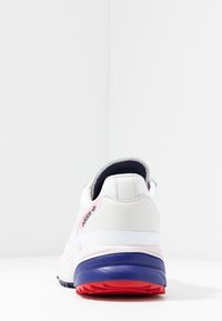 Vit sportskor med en texturerad yta, utrustad med en rosa logotyp på hälen och en flerfärgad sula med röda och blå accenter.