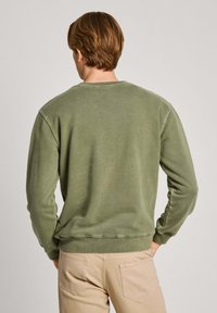 Hombre con cabello corto castaño, vistiendo un sudadera verde oliva y pantalones beige, de pie de espaldas con las manos en los bolsillos frente a un fondo plano.