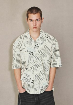 Young man with short hair wearing a newspaper print short-sleeve shirt and black pants, standing with hands in pockets against plain background.