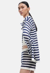 Raidallinen neulekortti tummansinisessä ja valkoisessa, jossa on avointadonkeli muotoilu, istuva vartalo ja sahalaitainen helma, yhdistettynä vastaaviin shortseihin.