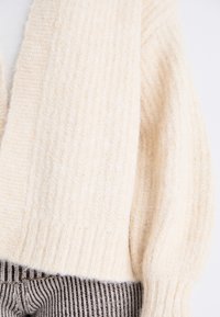 Cream-colored knitted cardigan with a ribbed texture and a slightly oversized fit, featuring long sleeves and a open-front design.
