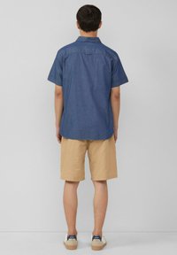 Camicia in chambray blu scuro a maniche corte con orlo arrotondato, abbinata a pantaloni corti beige chiaro e sneakers bianche con dettagli blu navy.