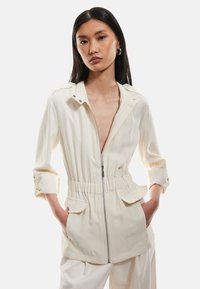 Giacca zip-up color crema con vita elastica, due tasche frontali e maniche arrotolate. Presenta dettagli con bottoni a scatto e colletto.