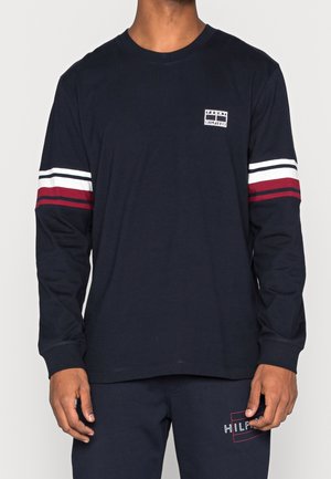 Mężczyzna ubrany w granatową koszulę z długim rękawem, z białymi i czerwonymi paskami na rękawach oraz małym logo na piersi, w połączeniu z pasującymi granatowymi spodniami.