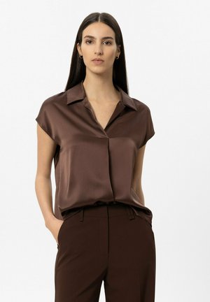 Frau mit langem dunklem Haar, die eine braune Satinbluse mit kurzen Ärmeln und passende braune Hose trägt, steht mit den Händen in den Taschen vor weißem Hintergrund.