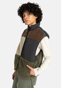 Een persoon met krullend haar poseert in een multi-kleurige vest. Het vest heeft verschillende texturen in aardetinten, met een stijlvolle, moderne snit.