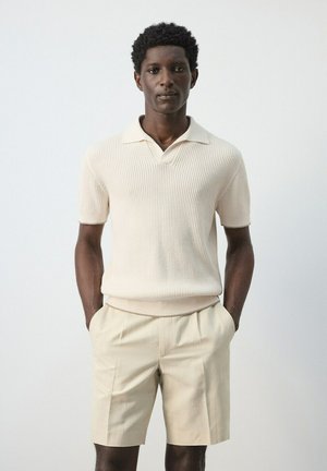 Joven de pie con las manos en los bolsillos, vistiendo un polo de punto de manga corta de color crema y pantalones cortos beige, contra un fondo claro y liso.