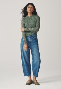 Green floral-patterned long-sleeve top with a fitted, smocked design, paired with loose blue denim jeans and olive green flat shoes.