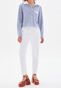 Camicia a righe blu e bianca, abbinata a pantaloni bianchi aderenti e sandali con zeppa marroni a strisce. Dettaglio della tasca sul petto molto interessante.