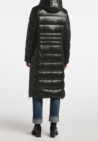 Osoba v dlouhém černém prošívaném péřovém kabátu s kapucí, s ohrnutými modrými džíny a čern�ými botami na podpatku, otočená zády na bílém pozadí.