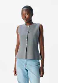 Haut sans manches gris avec un col rond, présentant une fermeture à boutons, un ourlet peplum et une texture lisse. Associé avec un jean bleu clair.