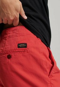 Pantalones cortos de algodón rojos con un bolsillo trasero visible y una etiqueta negra. La tela tiene una ligera textura y un botón marrón resalta el bolsillo.
