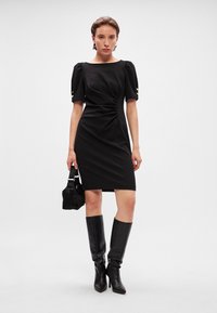 Crna uska haljina s puf rukavima, suptilnim nabiranjem oko struka i akcentima zlatnih dugmadi. Uparena s crnim čizmama do koljena i malom torbicom.