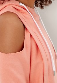 Sweatshirt léger en corail avec découpes aux épaules, capuche à cordon de serrage et cordon blanc. Texture douce, présentant des bords cousus.