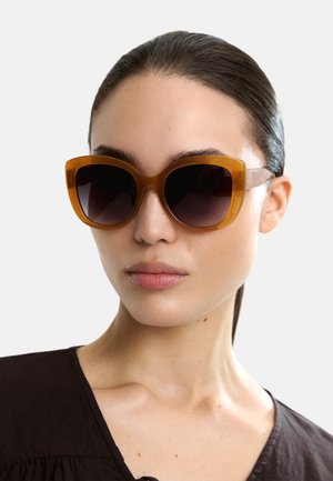 Pomarańczowe, oversize, kocie okulary przeciwsłoneczne z ciemnymi gradientowymi soczewkami i przezroczystymi zausznikami.