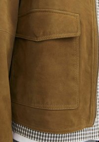 Brown suede jacket featuring a large patch pocket, contrasting stitching, and a smooth texture, layered over a checked shirt.