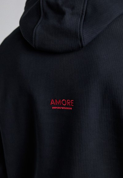 Hanorac negru cu model texturat, având un logo brodat roșu, "AMORE EMPORIOARMANI," amplasat pe partea superioară a spatelui. Textură moale, glugă ajustabilă.