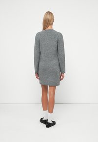 Γκρι πλεκτό φόρεμα με μακριά μανίκια, προσαρμοσμένη σιλουέτα και ριμπ τελείωμα. Φοριέται με μαύρα παπούτσια και άσπρες κάλτσες, παρουσιάζοντας έναν απλό, κλασικό σχεδιασμό.