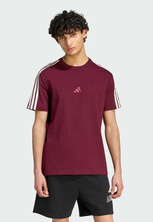 Ung mand med krøllet hår iført en burgundy Adidas t-shirt med hvide striber på ærmerne og sorte Adidas shorts, står mod en ensfarvet baggrund.