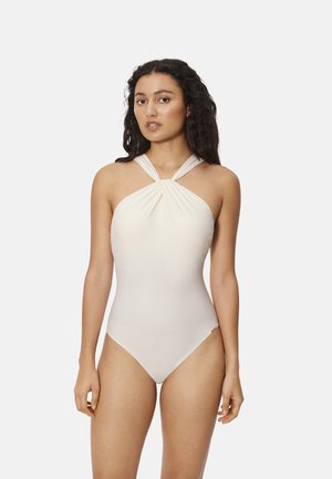 Mujer con cabello largo y rizado, vistiendo un traje de baño de una pieza color crema sin mangas, con cuello halter, sobre un fondo liso.