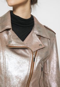 Błyszcząca, metaliczna różowa kurtka motocyklowa z suwakiem z przodu, srebrnymi elementami i pagonami; noszona na czarnym golfie.