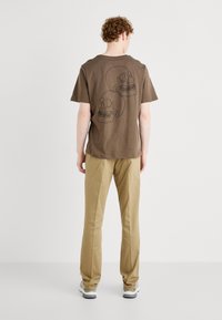Brown cotton t-shirt featuring a graphic of two skulls on the back. Paired with beige pants and grey sneakers, viewed from the back.